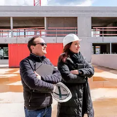 Zwei Personen mit Helm und Unterlagen bei der Baustellenbegehung des neuen Firmengebäudes der Sutter Medizintechnik GmbH in Emmendingen. Im Hintergrund ist der Rohbau mit Betonfassade zu sehen.