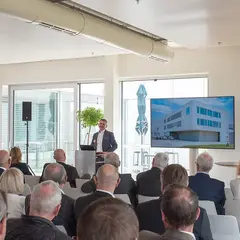 Bert Sutter spricht vor Publikum bei einer Veranstaltung zum Neubau der Sutter Medizintechnik GmbH, im Hintergrund ist ein Bildschirm mit der Visualisierung des neuen Gebäudes zu sehen.