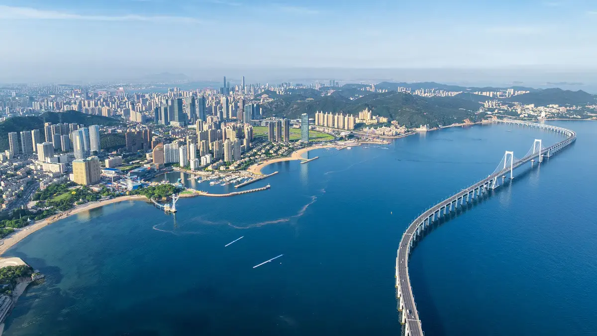 Panoramaaufnahme der Küstenstadt Dalian in China bei Abenddämmerung mit Hochhäusern, beleuchteter Uferpromenade und ruhigem Meer.