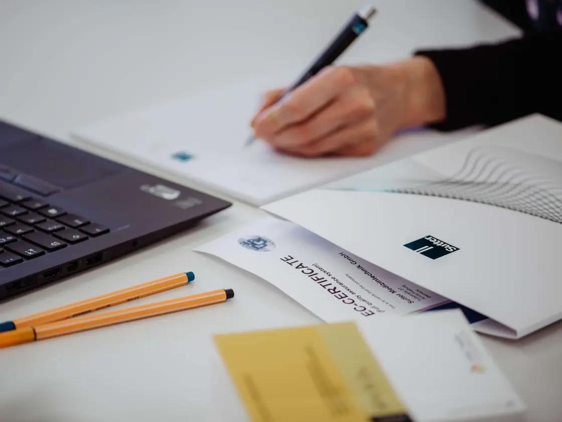 Nahaufnahme eines Schreibtisches mit Laptop, Bewerbungsunterlagen und Hand beim Schreiben – Symbolbild für den Bewerbungsprozess bei Sutter Medizintechnik