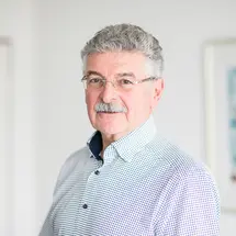 Dr. Zavisic mit Brille, grauem Haar und kariertem Hemd vor hellem Hintergrund.