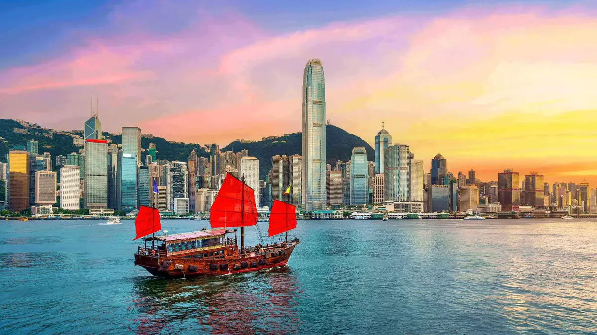 Traditionelles chinesisches Junkboot mit roten Segeln fährt vor der Skyline von Hongkong bei farbenprächtigem Sonnenuntergang über das Wasser.        