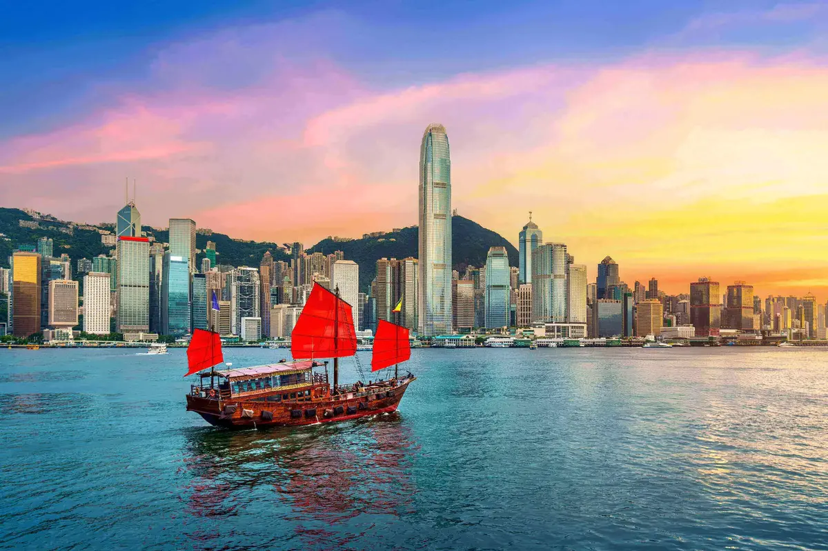 Traditionelles chinesisches Junkboot mit roten Segeln fährt vor der Skyline von Hongkong bei farbenprächtigem Sonnenuntergang über das Wasser.        