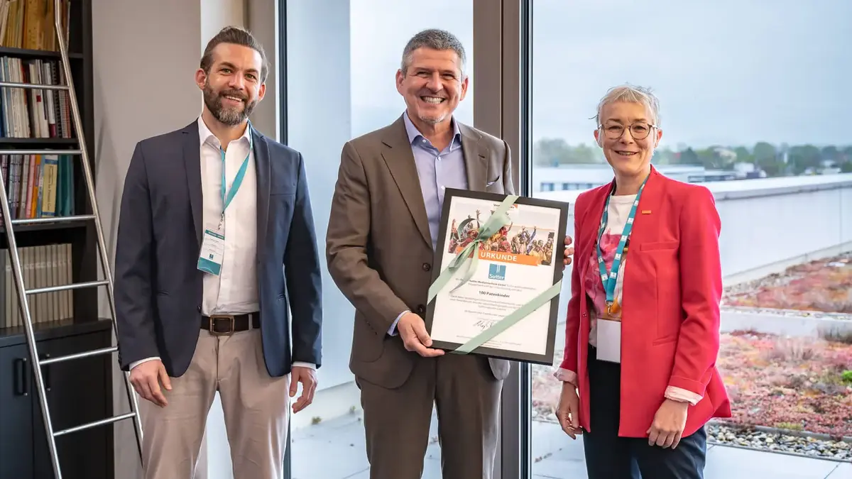 Drei Personen stehen lächelnd zusammen; in der Mitte wird eine gerahmte Urkunde von World Vision mit einer grünen Schleife überreicht, die 100 Patenschaften würdigt; im Hintergrund Büro mit Bücherregal und Fensterfront.