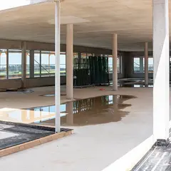 Blick in den Innenausbau des Sutter-Neubaus in Emmendingen mit großen Fensterflächen, Betonstützen und abgestellten Glasscheiben. Pfützen auf dem Boden spiegeln das Tageslicht.