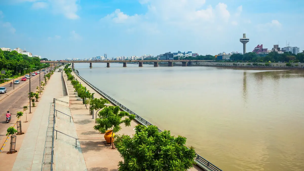 Ahmedabad riverfront with bridge, trees, promenade, and modern city skyline.