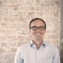 Person in hellblauem Hemd lächelnd vor einer Backsteinwand