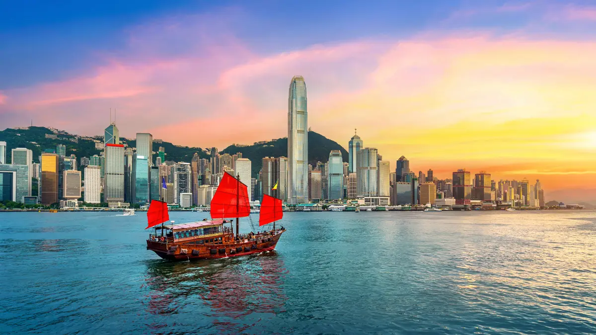 Traditionelles chinesisches Junkboot mit roten Segeln fährt vor der Skyline von Hongkong bei farbenprächtigem Sonnenuntergang über das Wasser.        