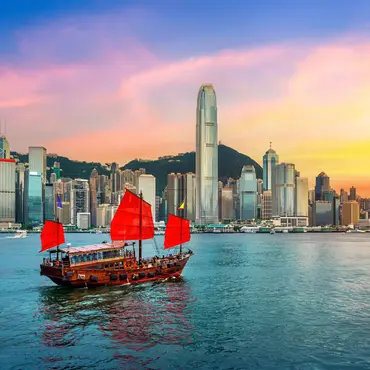 Traditionelles chinesisches Junkboot mit roten Segeln fährt vor der Skyline von Hongkong bei farbenprächtigem Sonnenuntergang über das Wasser.        