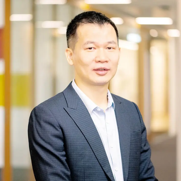 Eine Person in dunklem Sakko und hellblauem Hemd steht in einem Büroflur mit Glaswänden und Holzverkleidung. Im Hintergrund sind unscharfe Büroleuchten und Türen zu sehen.