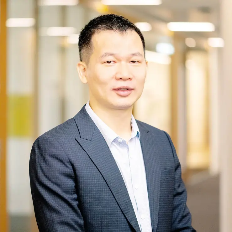 Eine Person in dunklem Sakko und hellblauem Hemd steht in einem Büroflur mit Glaswänden und Holzverkleidung. Im Hintergrund sind unscharfe Büroleuchten und Türen zu sehen.