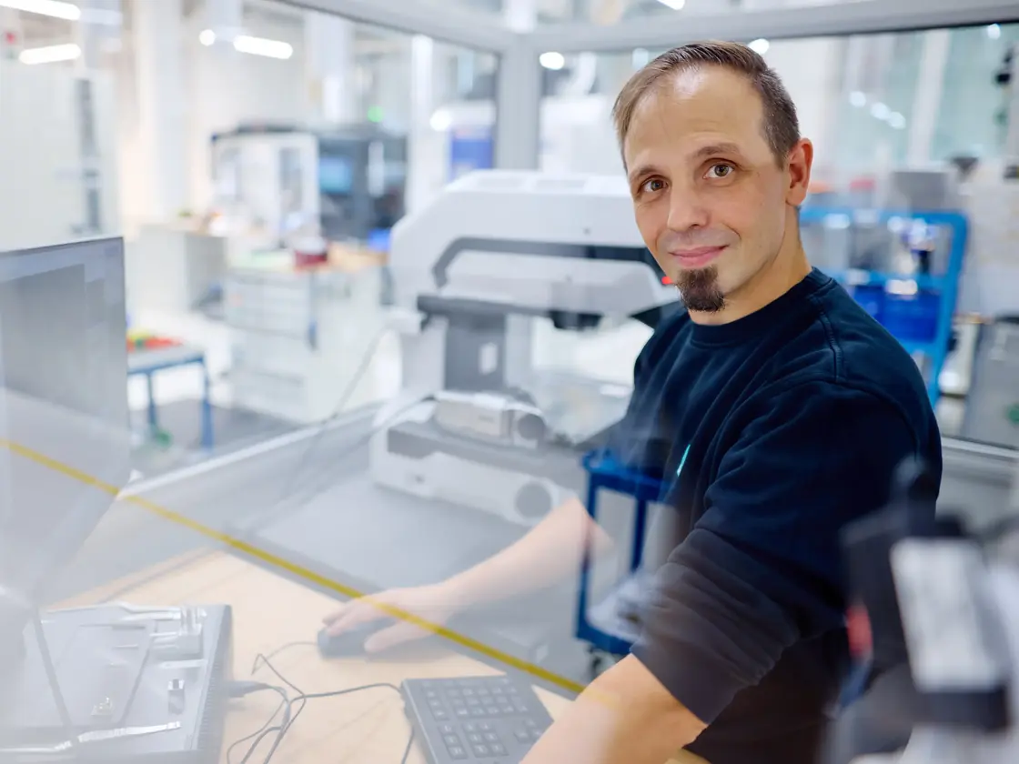 Mitarbeiter von Sutter Medizintechnik steht an einem Arbeitsplatz in der CNC und blickt lächelnd in die Kamera. Im Hintergrund sind moderne Prüfgeräte und Fertigungsbereiche zu sehen.